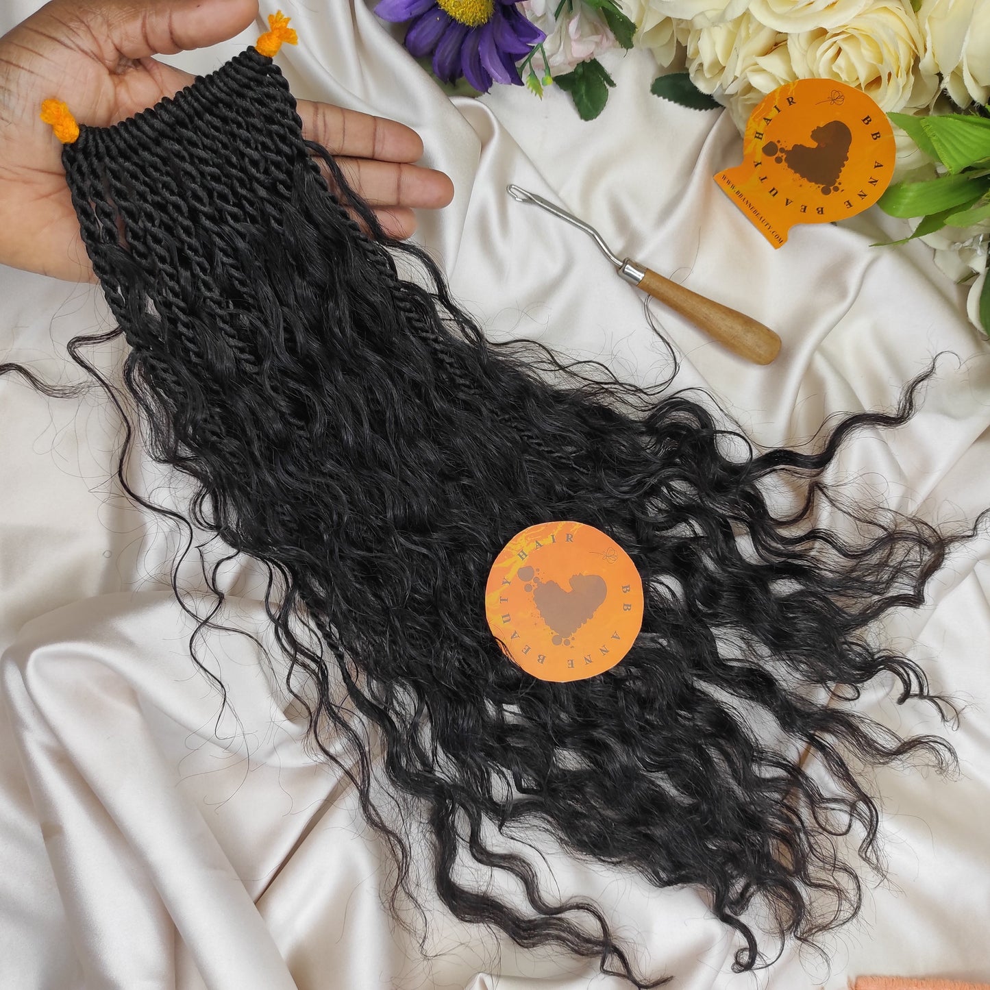 Black braided hair extensions held by a hand on a white fabric background with floral decorations.