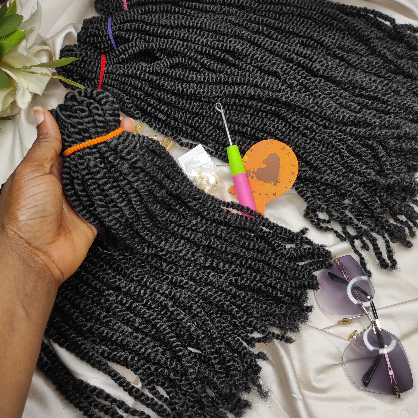 Black braided hair extensions with a hand holding a section, on a white surface with sunglasses and a heart-shaped object.