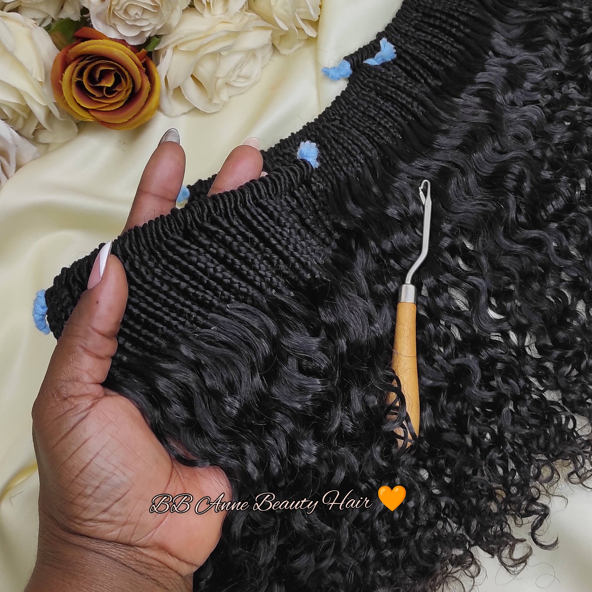 Black curly hair extensions held by a hand with a crochet hook on a floral background, featuring B.B.