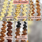 A hair color chart showing six wavy hair extensions in different shades, labeled Col 613, Col 27/613, Col 30/613, Col 33/30, and Col B29 (Ombre 1/30). The chart displays light yellow, blonde, light brown, medium brown, dark brown, and black ombre styles against a gray textured background.