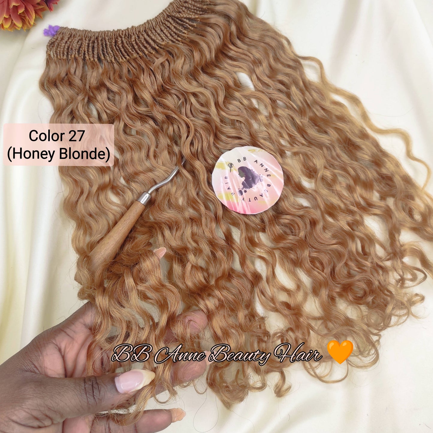 A bundle of wavy, curly hair extensions in Honey Blonde color, held by a hand over a cream-colored fabric backdrop with decorative flowers and a small tool.