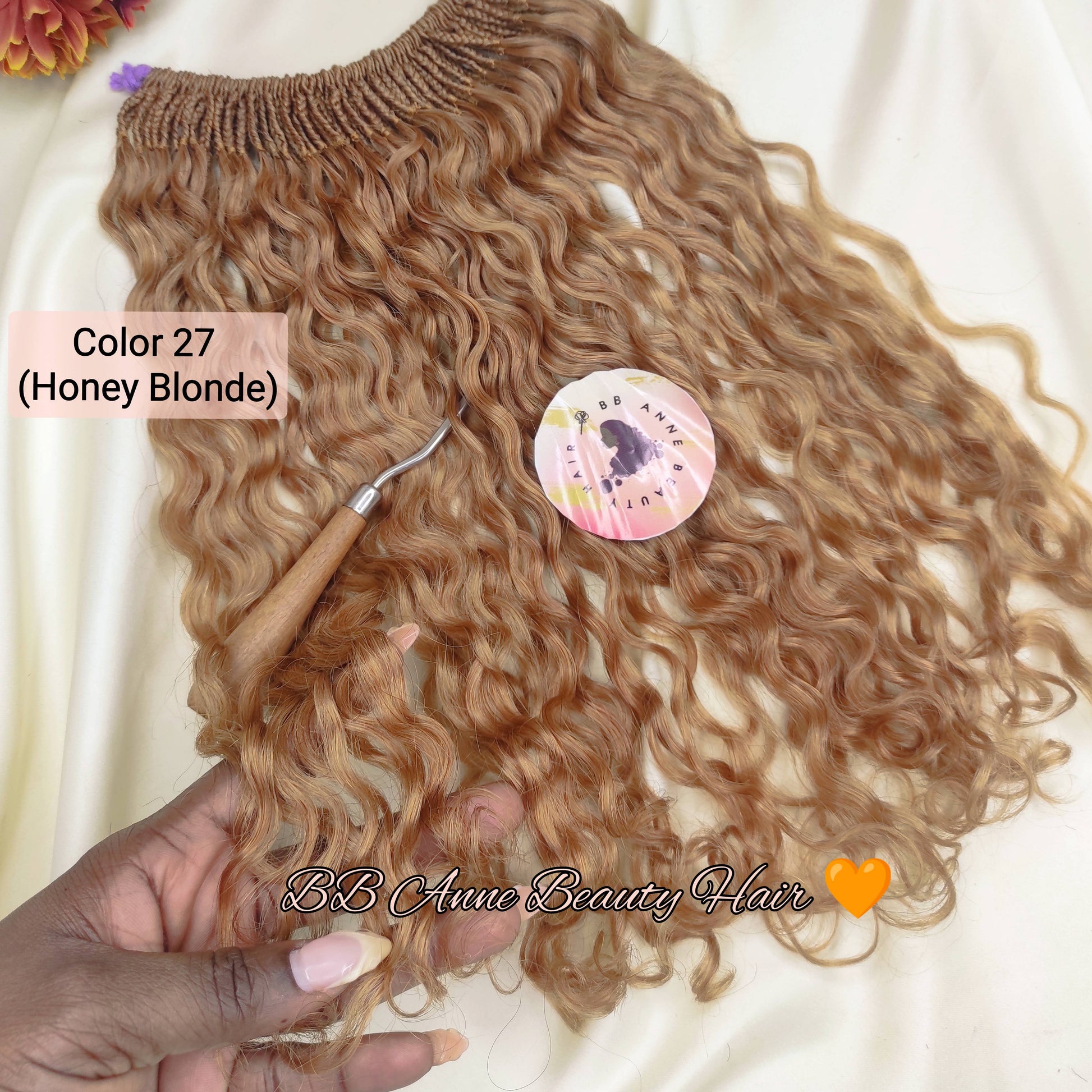 A bundle of wavy, curly hair extensions in Honey Blonde color, held by a hand over a cream-colored fabric backdrop with decorative flowers and a small tool.