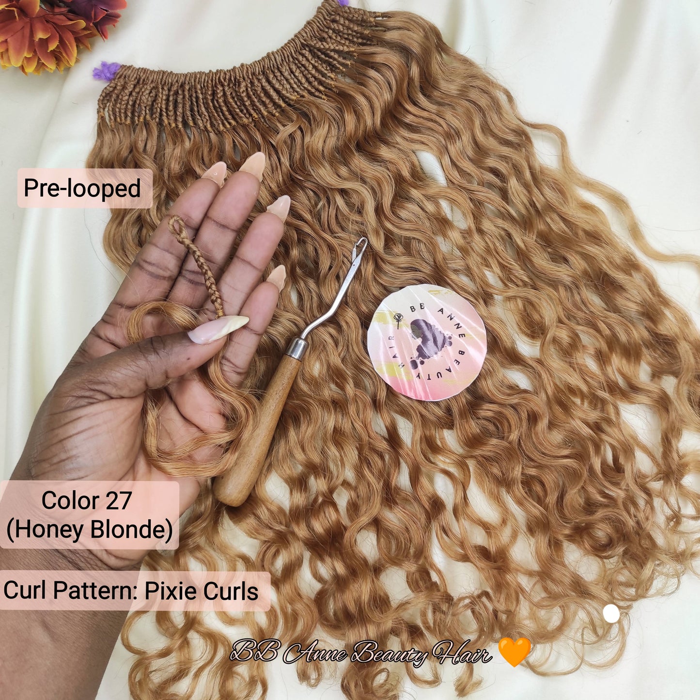 A hand holds a pre-looped box braiding hair extension with tight, glossy curly locks in a honey blonde shade. The hair is displayed on a white fabric background with decorative flowers, a crochet hook tool, and text labels indicating 'Pre-looped', 'Color 27 (Honey Blonde)', and 'Curl Pattern: Pixie Curls'.