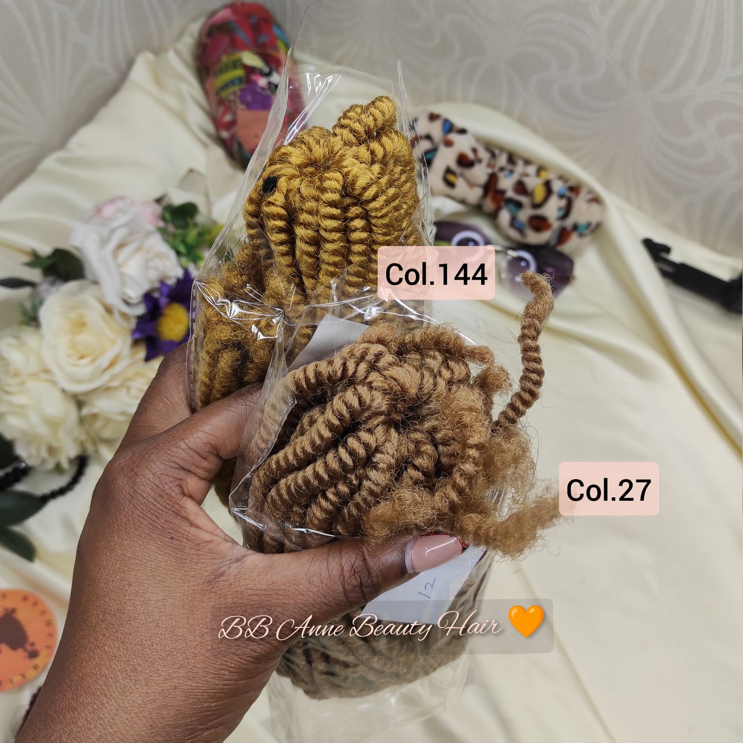 A hand holds two packaged bundles of coiled curly hair extensions in different shades: a golden blonde/brown color labeled Col.144 and a light brown color labeled Col.