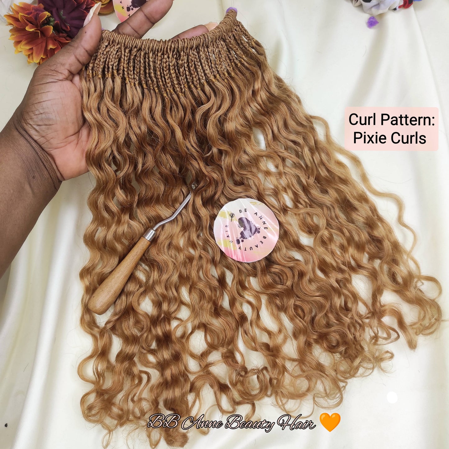 A hand holds a bundle of long, curly hair extensions with a textured elastic top, styled with a curling iron and a round hair accessory on a white fabric background with decorative flowers.