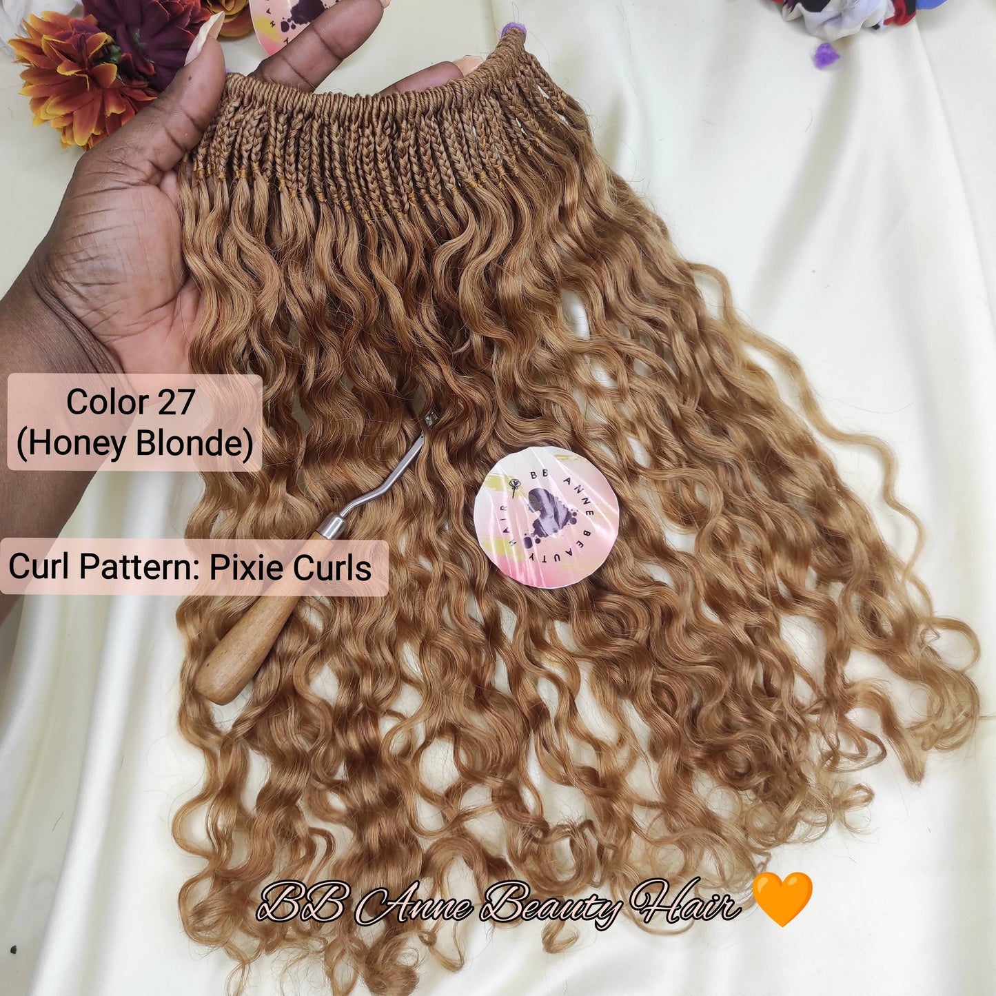 A hand holds a bundle of curly hair extensions in honey blonde color against a white fabric background with decorative flowers. Text labels indicate Color 27 (Honey Blonde) and Curl Pattern: Pixie Curls.