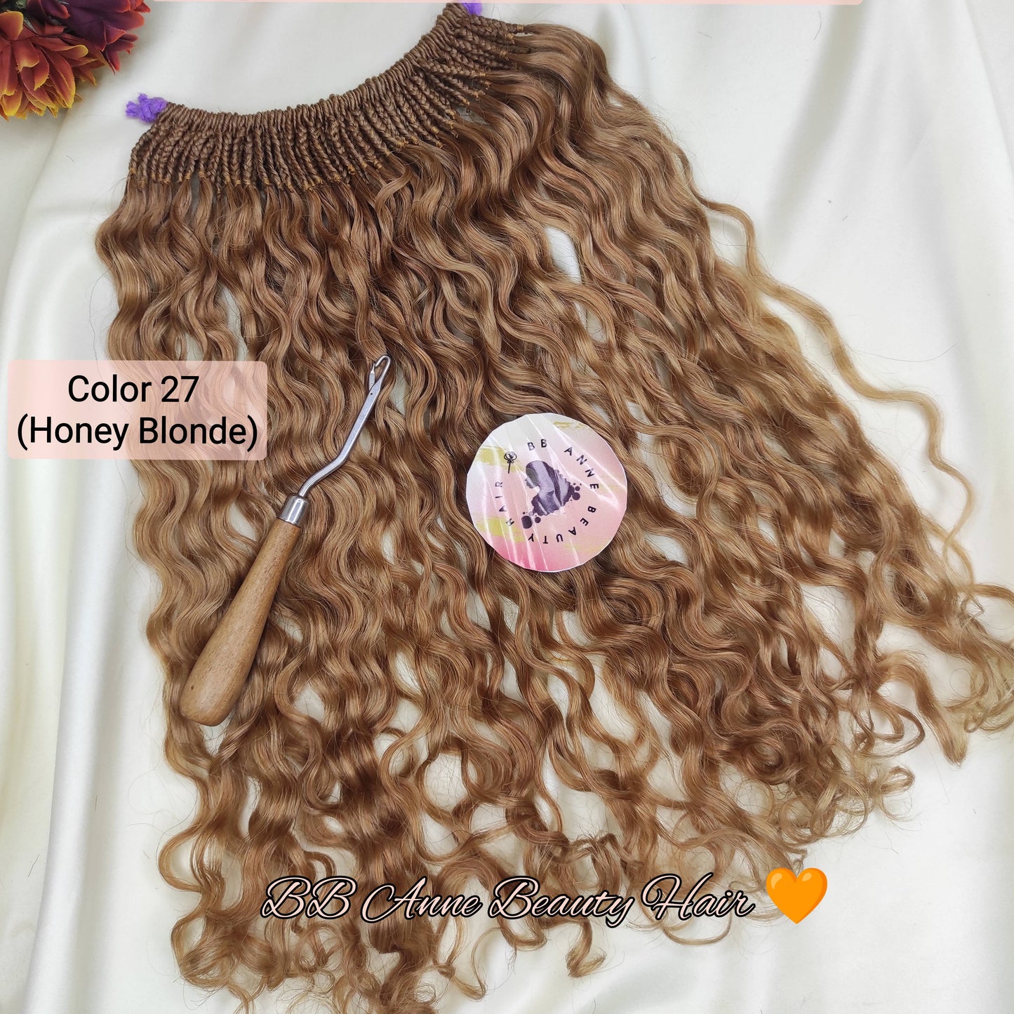 A bundle of pre-braided human hair crochet braids in a honey blonde shade, labeled 'AMARA' and 'Color 27 (Honey Blonde)', displayed on a white fabric background with a styling tool and decorative flowers.