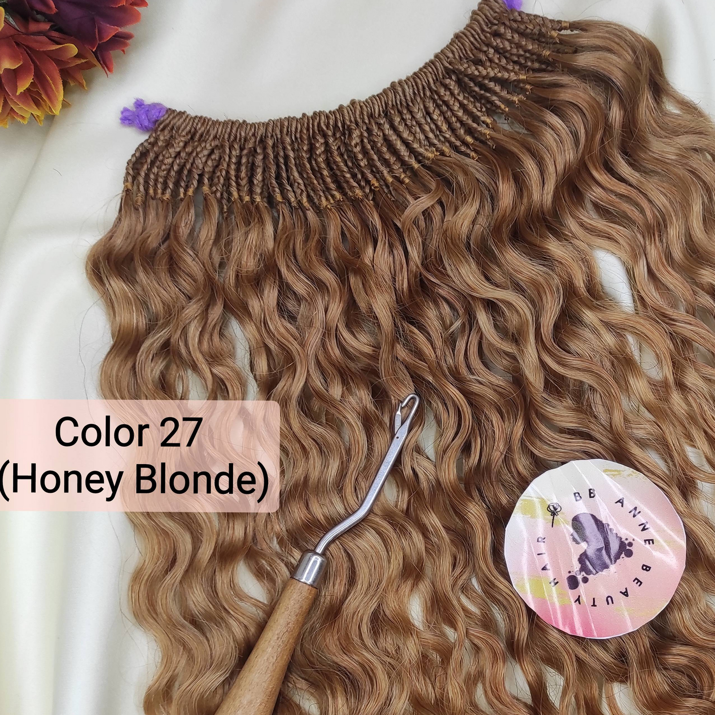 A bundle of pre-braided human hair crochet braids in a honey blonde shade, labeled 'AMARA' and 'Color 27 (Honey Blonde)', displayed on a white fabric background with a styling tool and decorative flowers.