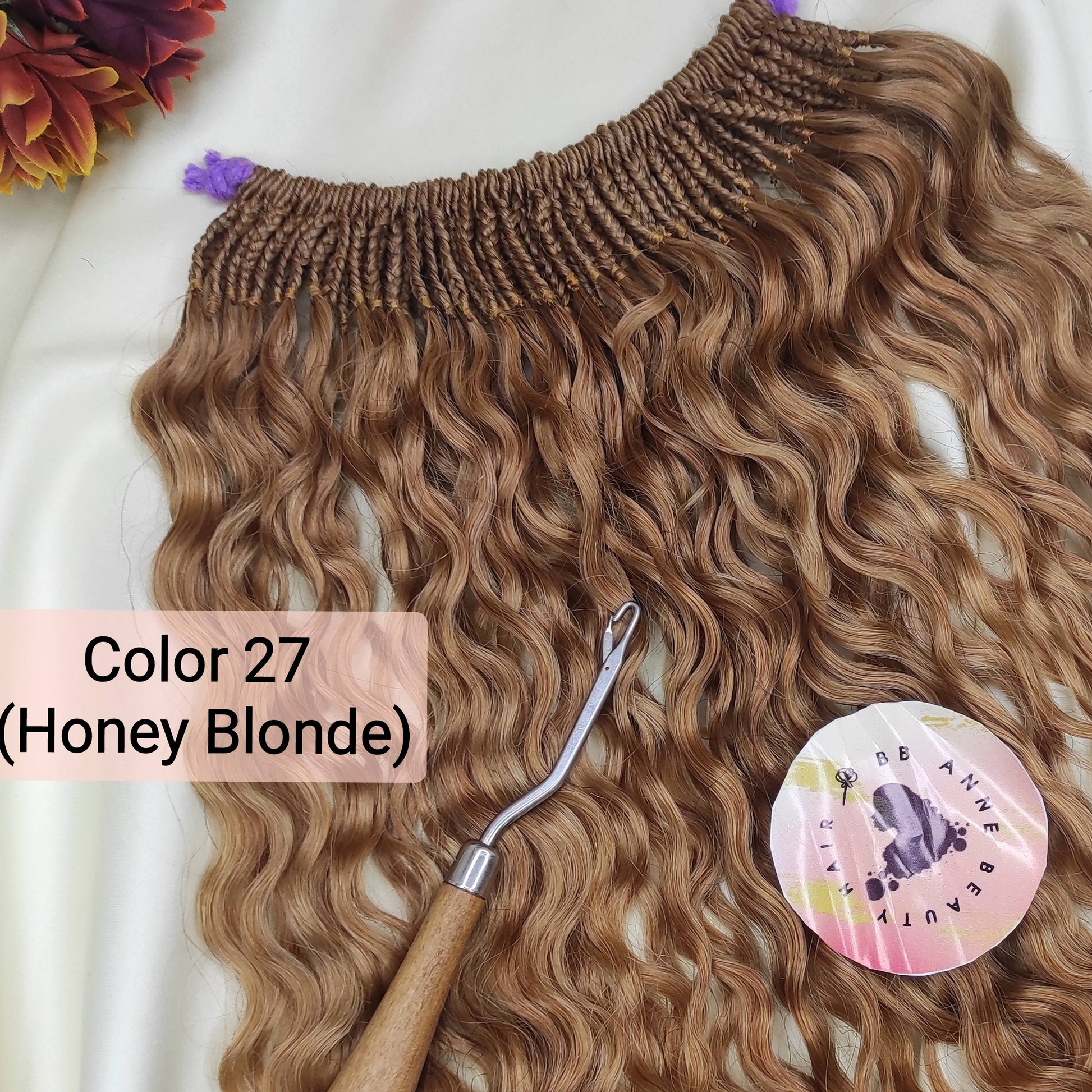 A bundle of pre-braided human hair crochet braids in a honey blonde shade, labeled 'AMARA' and 'Color 27 (Honey Blonde)', displayed on a white fabric background with a styling tool and decorative flowers.